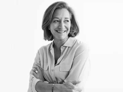 Monochrome portrait of Fran Horowitz standing against a clean white background with her arms crossed and a warm smile. She wears a light button-up blouse with chest pockets, minimal jewelry, and a wristwatch, while her short, softly styled hair frames her face. The high-key black-and-white composition creates a polished, professional look that emphasizes confidence, approachability, and leadership