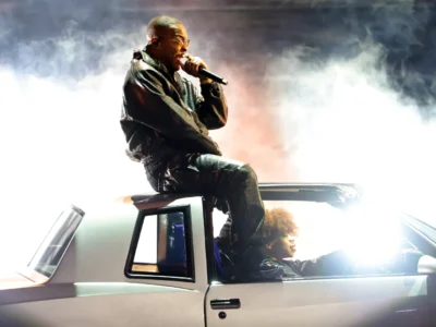 T.I. sits on the roof of a white sports car while singing into a microphone, surrounded by dramatic stage smoke and bright backlighting as another person sits behind the wheel during a live perform