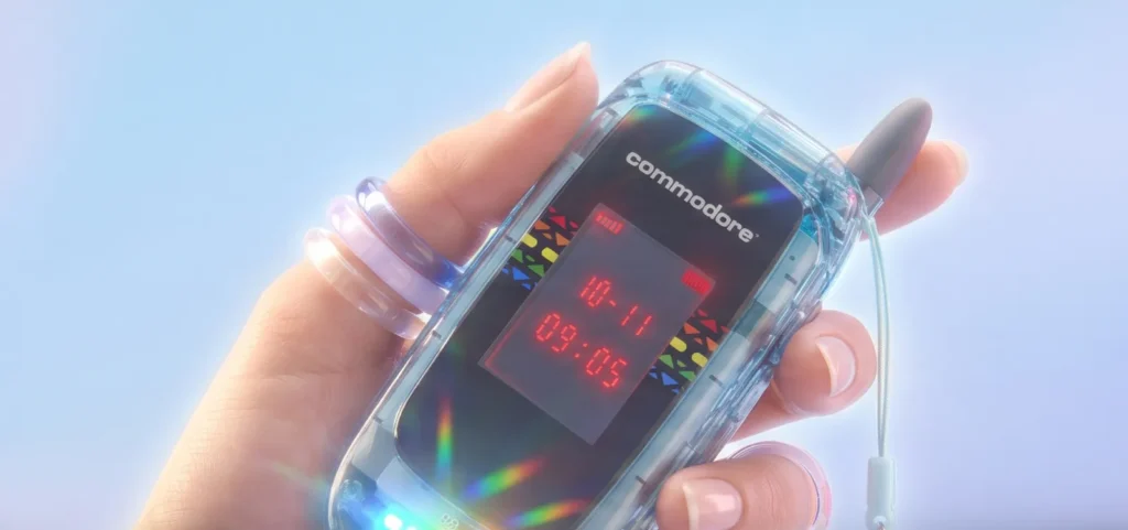 A translucent blue Commodore flip phone is held in one hand, highlighting its transparent shell, external LED-style display, rainbow graphics, illuminated accents, and antenna-inspired detail. The colorful design evokes late-1990s consumer electronics while incorporating modern materials and finish