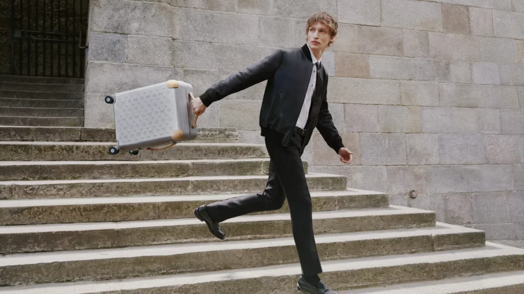 Model in a black leather bomber jacket and pinstripe trousers strides down stone steps while carrying a silver Louis Vuitton Horizon aluminum suitcase embossed with the Monogram pattern