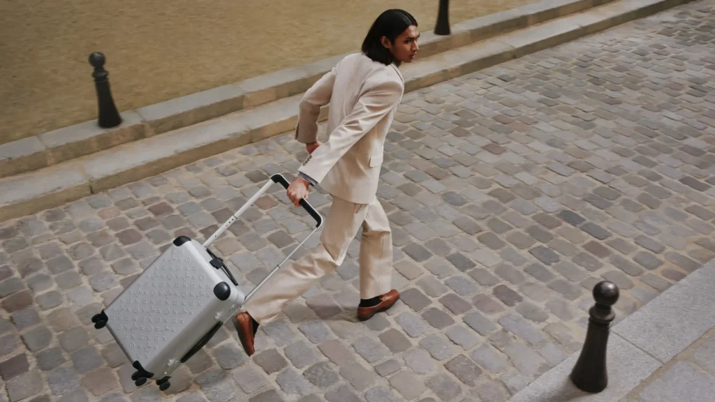 Model in a light beige tailored suit moves across a cobblestone street while pulling a silver Louis Vuitton Horizon rolling suitcase with extended telescopic handle