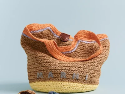 A woven Marni raffia tote in natural brown with pastel yellow and orange accents. Lavender trim outlines the opening, while detachable crocheted charms rest beside the bag, emphasizing its artisanal summer aesthetic