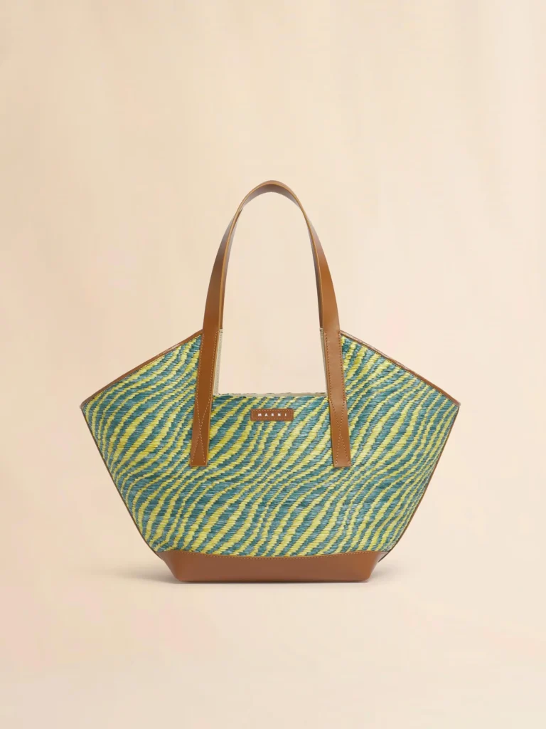 A geometric Marni tote crafted from woven material in blue, green, and yellow wave-like stripes. Tan leather handles and trim provide structure, creating a bold yet refined warm-weather carryall