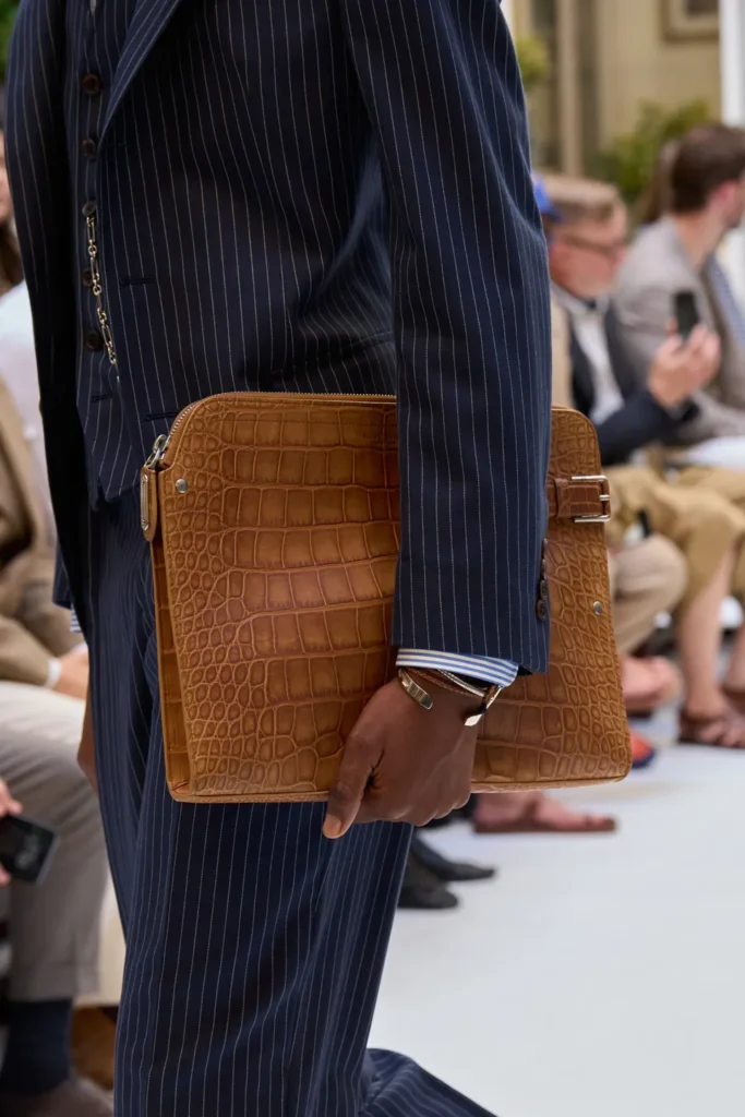 Close-up of a navy pinstripe double-breasted suit styled with a striped dress shirt, patterned tie, and oversized crocodile-embossed leather portfolio carried down the runway