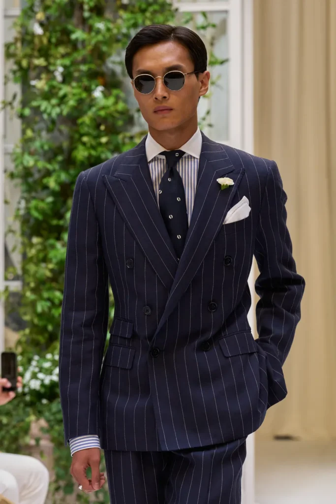 Model walking in a sharply tailored navy pinstripe suit featuring wide peak lapels, a striped shirt, patterned tie, white pocket square, and round sunglasses