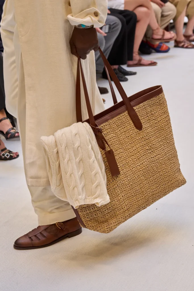 Natural raffia tote with brown leather trim and handles, paired with cream tailoring, woven leather sandals, and a draped cable-knit sweater
