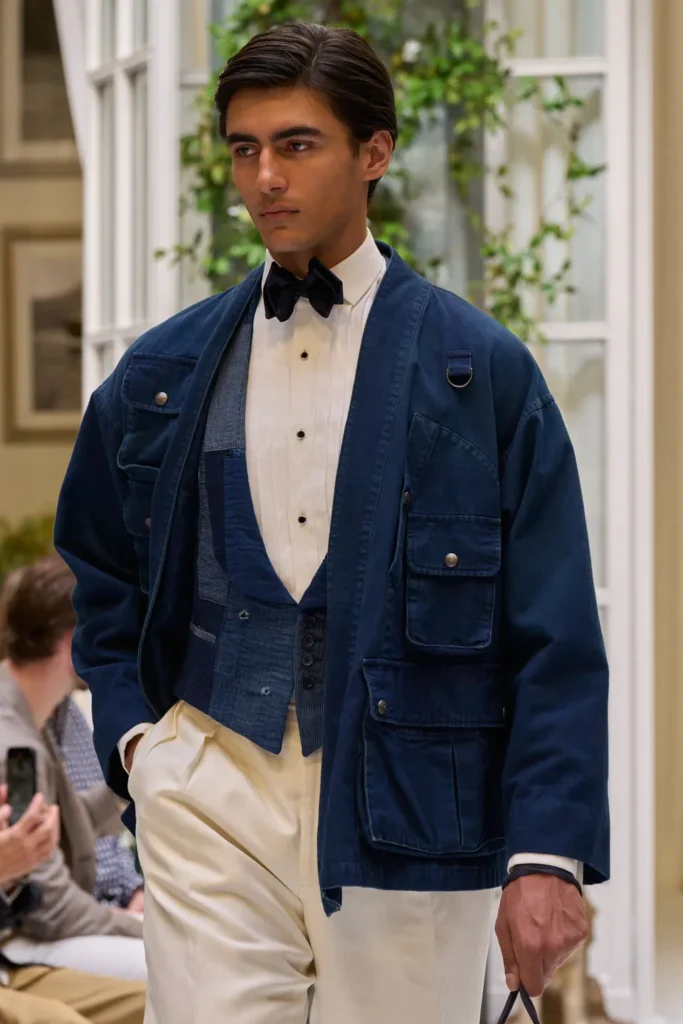 Layered tailoring look featuring a navy chore jacket over a patchwork waistcoat, white formal shirt, and cream pleated trousers
