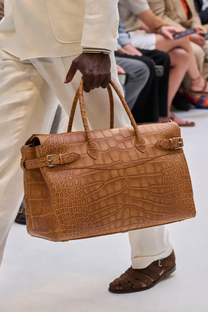 Large crocodile-embossed leather travel bag in warm tan carried with cream tailoring and woven leather fisherman sandal