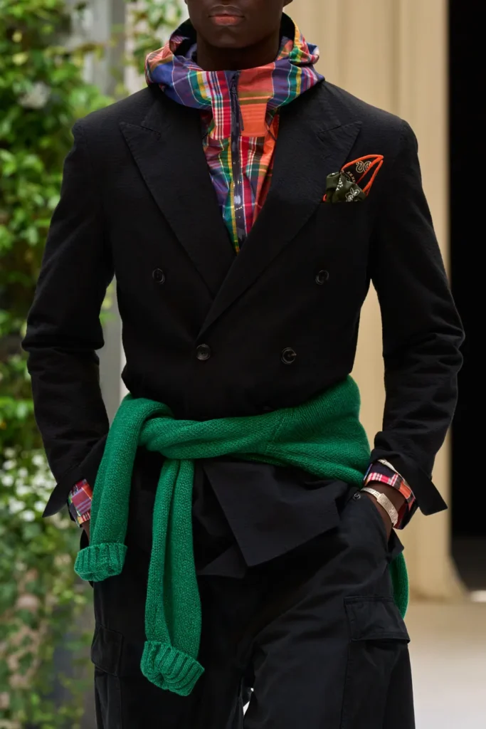 Modern tailored look combining a black double-breasted blazer, colorful plaid hooded layer, cargo trousers, and a bright green sweater tied at the waist