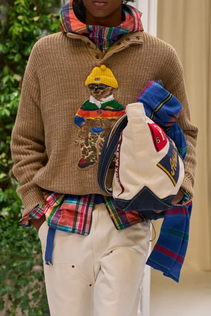 Chunky knit sweater featuring a signature bear graphic, layered over a vibrant plaid shirt and styled with a heritage canvas tote