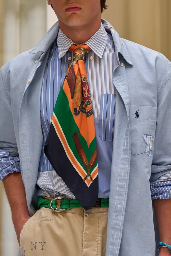 Light blue Oxford shirt layered over a striped button-down and colorful silk scarf, finished with distressed chinos and a bright green woven belt