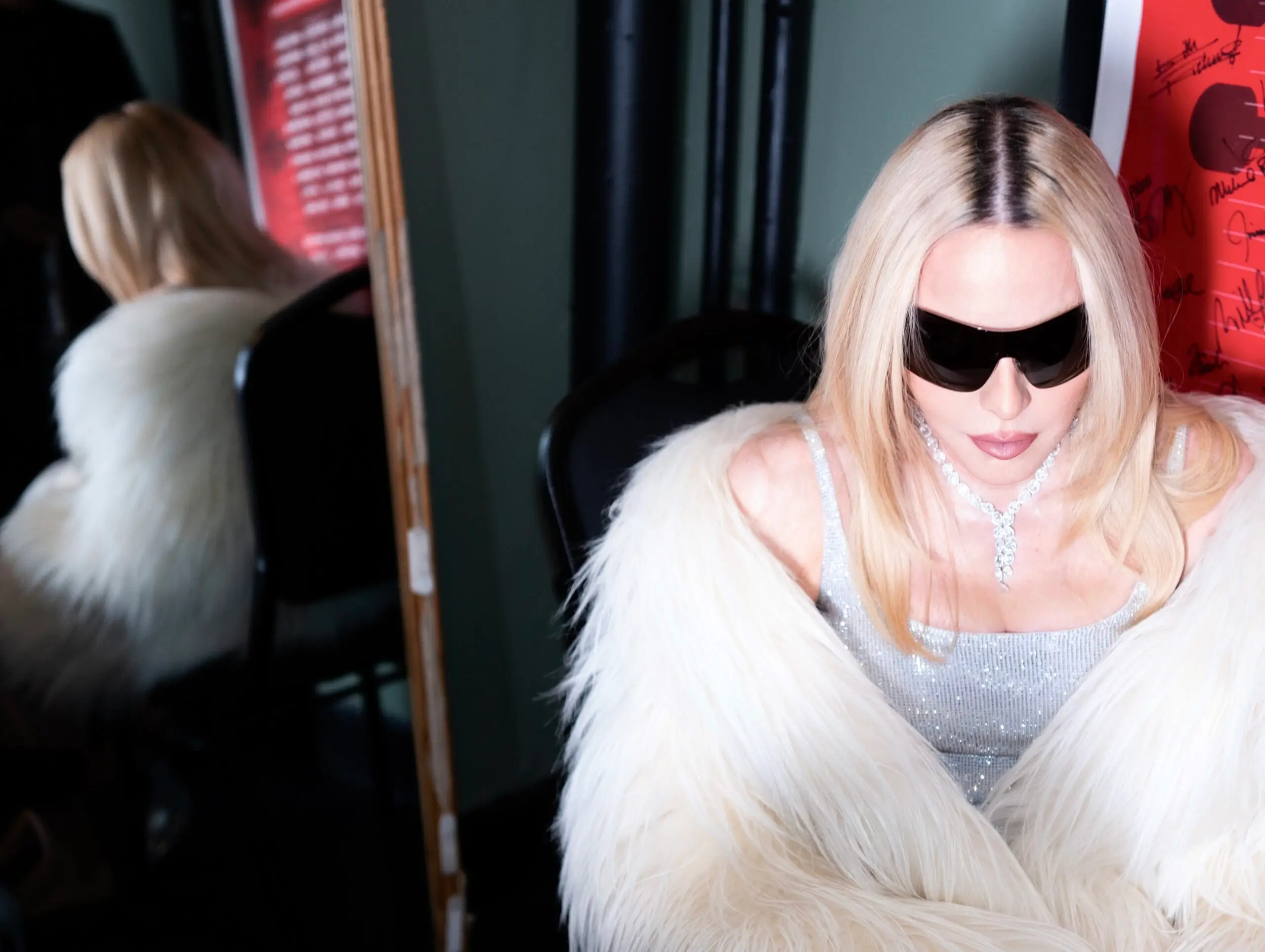 Portrait of a performer wearing oversized sunglasses, a crystal necklace, and a voluminous white faux-fur coat while seated backstage near framed memorabilia