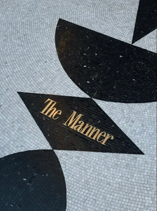 Black-and-white mosaic tile floor featuring geometric shapes and gold lettering spelling “The Manner,” creating an elegant Art Deco-inspired entrance detail