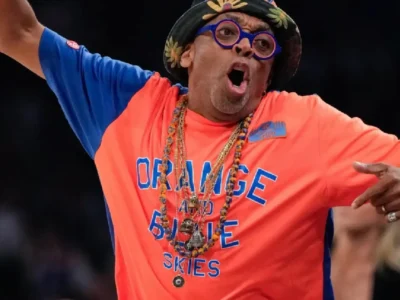Courtside sports fan dressed in a bright orange-and-blue outfit enthusiastically cheering during a basketball game. Spike Lee wears a floral bucket hat, round blue glasses, layered necklaces, and a graphic Tee reading “Orange and Blue Skies,” with arms spread wide in celebration. The arena background is softly blurred, emphasizing the energetic expression and vibrant team color