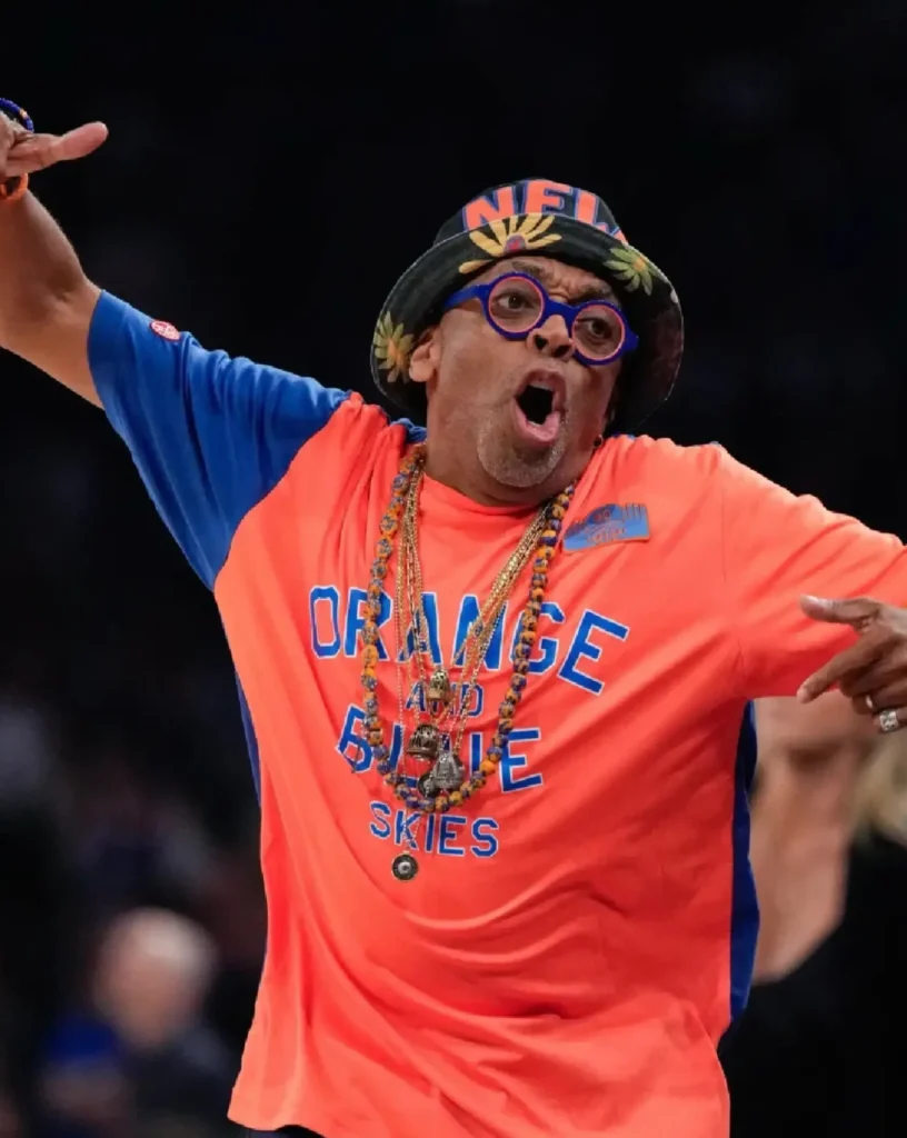 Courtside sports fan dressed in a bright orange-and-blue outfit enthusiastically cheering during a basketball game. Spike Lee wears a floral bucket hat, round blue glasses, layered necklaces, and a graphic Tee reading “Orange and Blue Skies,” with arms spread wide in celebration. The arena background is softly blurred, emphasizing the energetic expression and vibrant team color