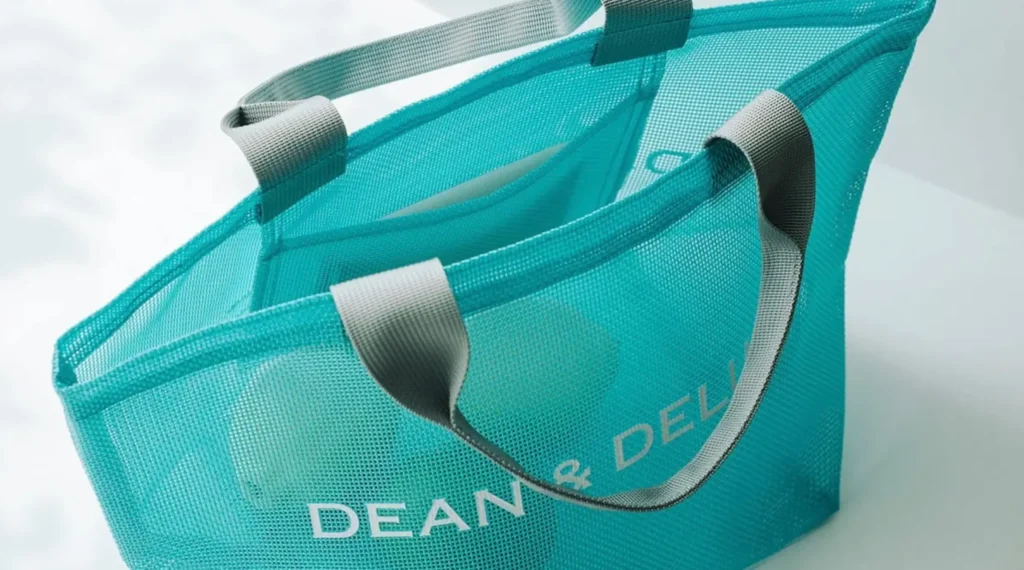 Close-up view of a turquoise DEAN & DELUCA mesh tote bag showcasing its semi-transparent construction, reinforced gray handles, interior pocket, and minimalist branding against a bright white background