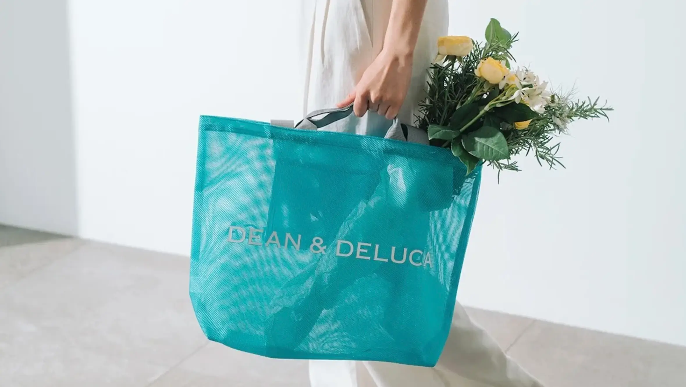 A person carrying a large turquoise DEAN & DELUCA mesh tote filled with fresh flowers. The translucent bag features gray handles and white logo branding, photographed in a bright, minimalist interior with soft natural light