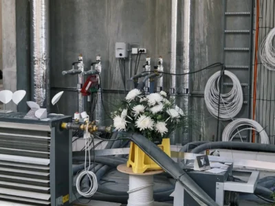 Industrial exhibition setup inside a raw concrete space, with exposed pipes, coiled cables, machinery, and a bouquet of white flowers arranged atop a cable spool, blending construction-site aesthetics with contemporary design presentation