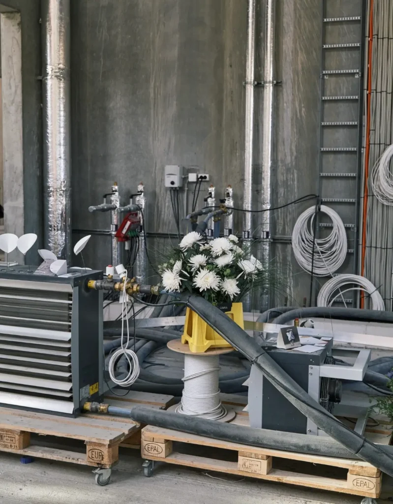 Industrial exhibition setup inside a raw concrete space, with exposed pipes, coiled cables, machinery, and a bouquet of white flowers arranged atop a cable spool, blending construction-site aesthetics with contemporary design presentation