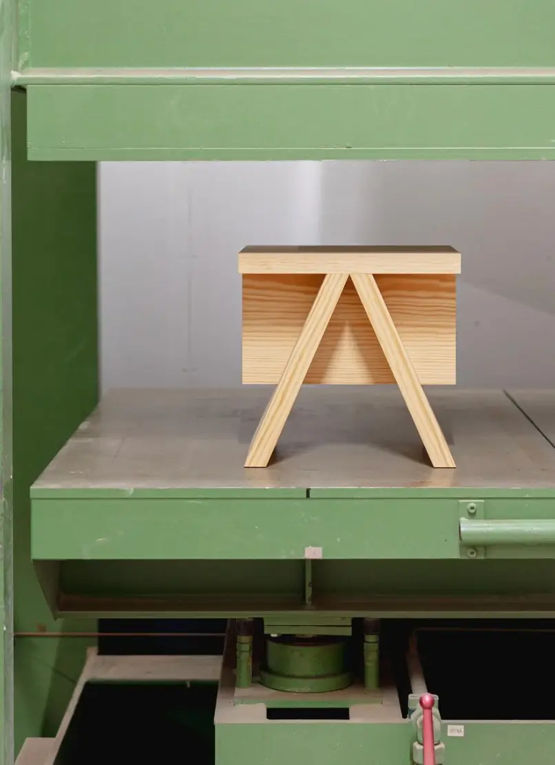 A handcrafted pinewood stool sits inside an industrial green press machine, highlighting its clean geometric form, natural wood grain, and minimalist design. The contrast between the warm timber and heavy machinery emphasizes the piece’s craftsmanship and production process