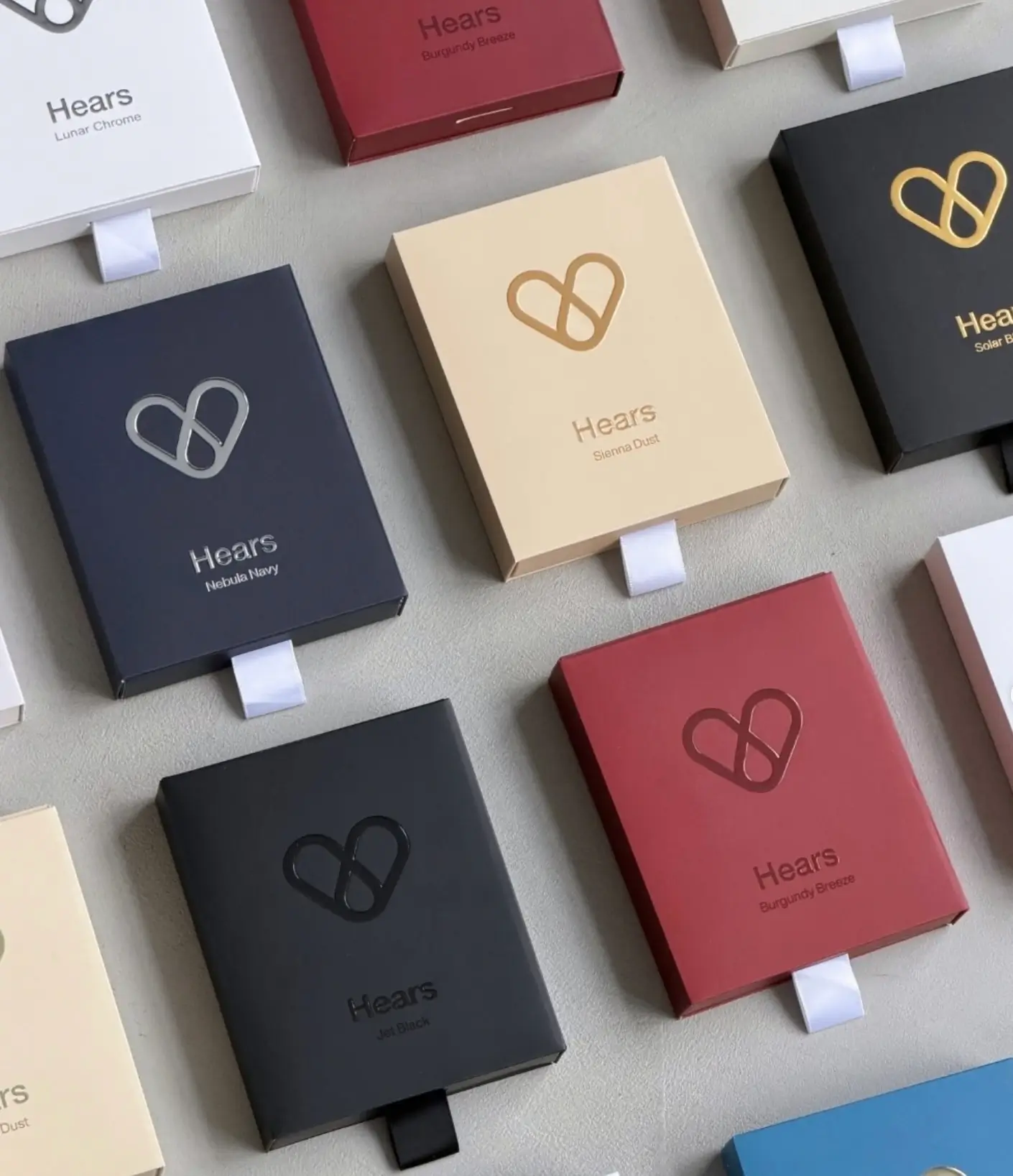 A second image presents a neatly arranged flat lay of Hears packaging in multiple colorways, including cream, navy, burgundy, black, and white. Each box features an embossed heart-shaped logo and color-specific product names such as Sierra Dust, Nebula Navy, Jet Black, Lunar Chrome, and Burgundy Breeze. The minimalist presentation highlights the brand’s refined packaging design, premium finishes, and cohesive visual identity through a palette of muted, contemporary tones