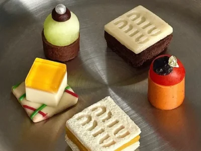 A curated assortment of miniature luxury-inspired desserts is arranged on a brushed stainless-steel tray. The sculptural pastries feature geometric forms, embossed branding details, vibrant glazes, and delicate garnishes, blending haute pâtisserie craftsmanship with fashion-house aesthetic