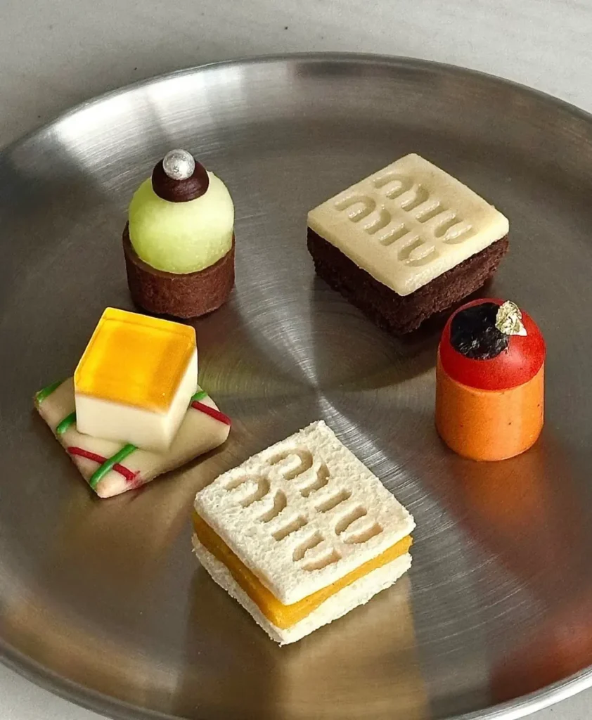 A curated assortment of miniature luxury-inspired desserts is arranged on a brushed stainless-steel tray. The sculptural pastries feature geometric forms, embossed branding details, vibrant glazes, and delicate garnishes, blending haute pâtisserie craftsmanship with fashion-house aesthetic