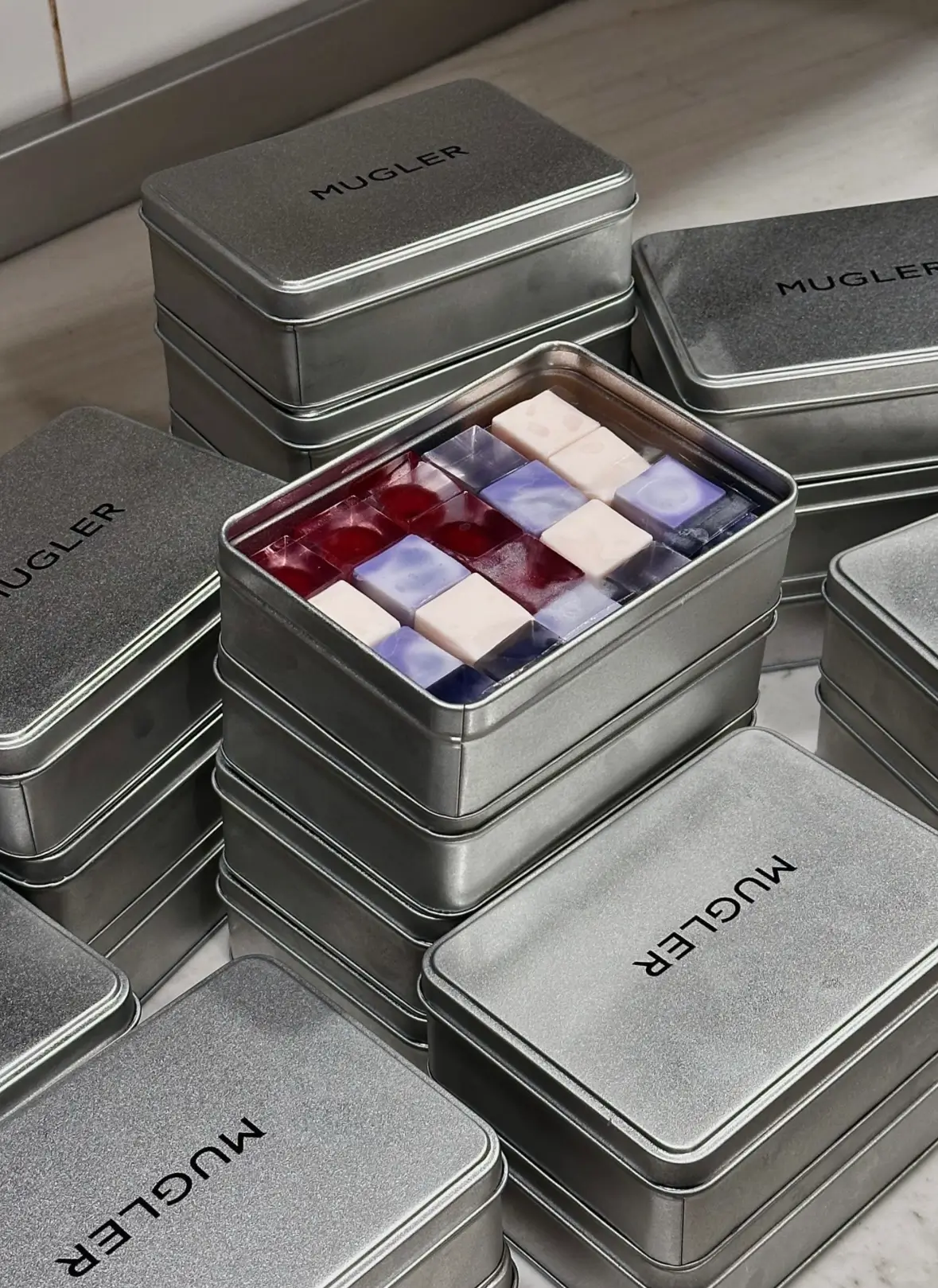 Branded metallic tins stamped with “MUGLER” are stacked across a marble surface, surrounding an open container filled with jewel-like confection cubes. The translucent sweets in shades of crimson, lavender, and ivory create a futuristic presentation that echoes the label’s bold visual identity