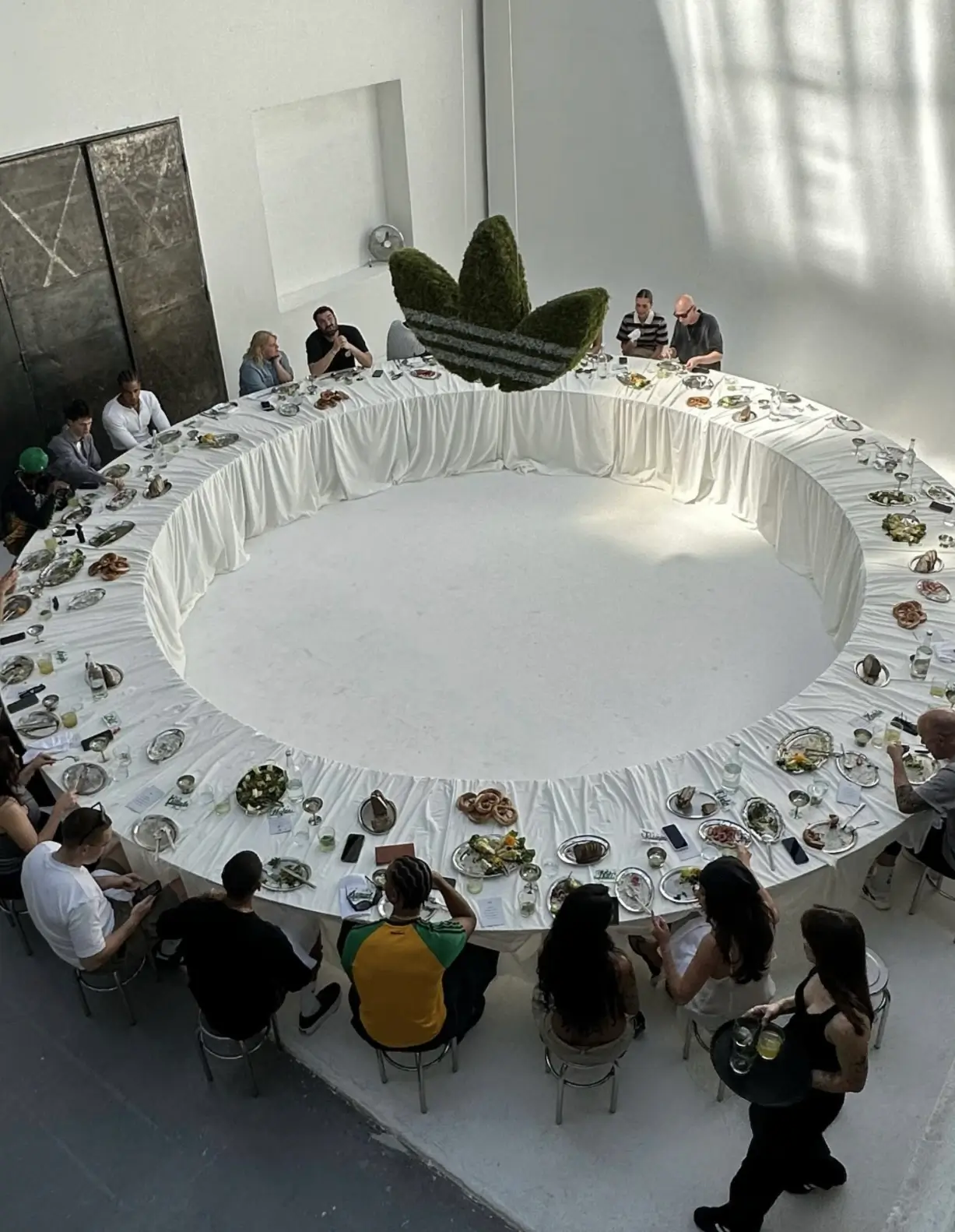 Guests gather around a dramatic circular dining installation draped in white linens inside a bright industrial space. Suspended above the center is a large moss-covered adidas Trefoil sculpture, transforming the communal meal into an immersive brand-led cultural experience