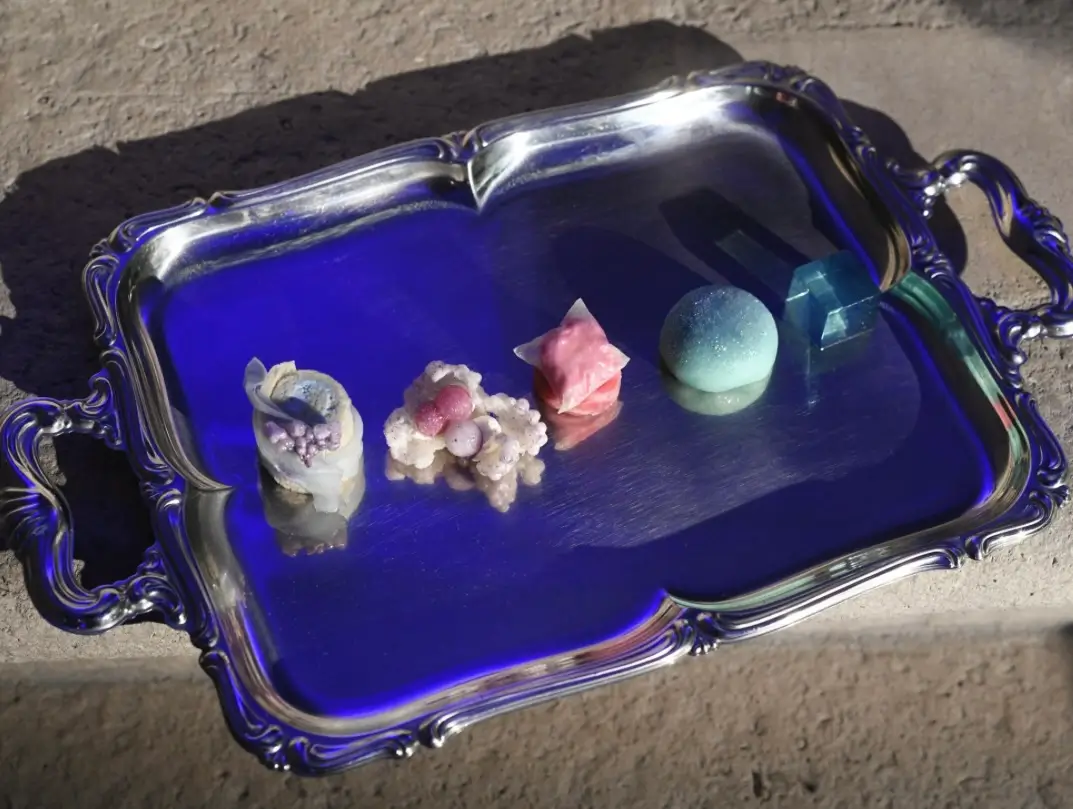 A silver serving tray bathed in cobalt-blue light displays a series of avant-garde bite-sized desserts resembling crystals, minerals, and abstract sculptures. The imaginative presentation combines fine dining, contemporary art, and fantasy-inspired design in a striking view composition