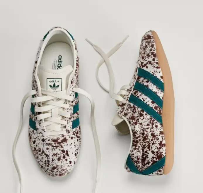 Top-down view of a pair of adidas low-profile sneakers featuring a white upper covered in brown speckled splatter detailing. Teal Three Stripes branding contrasts against the textured surface, while white laces and a gum sole complete the retro-inspired design
