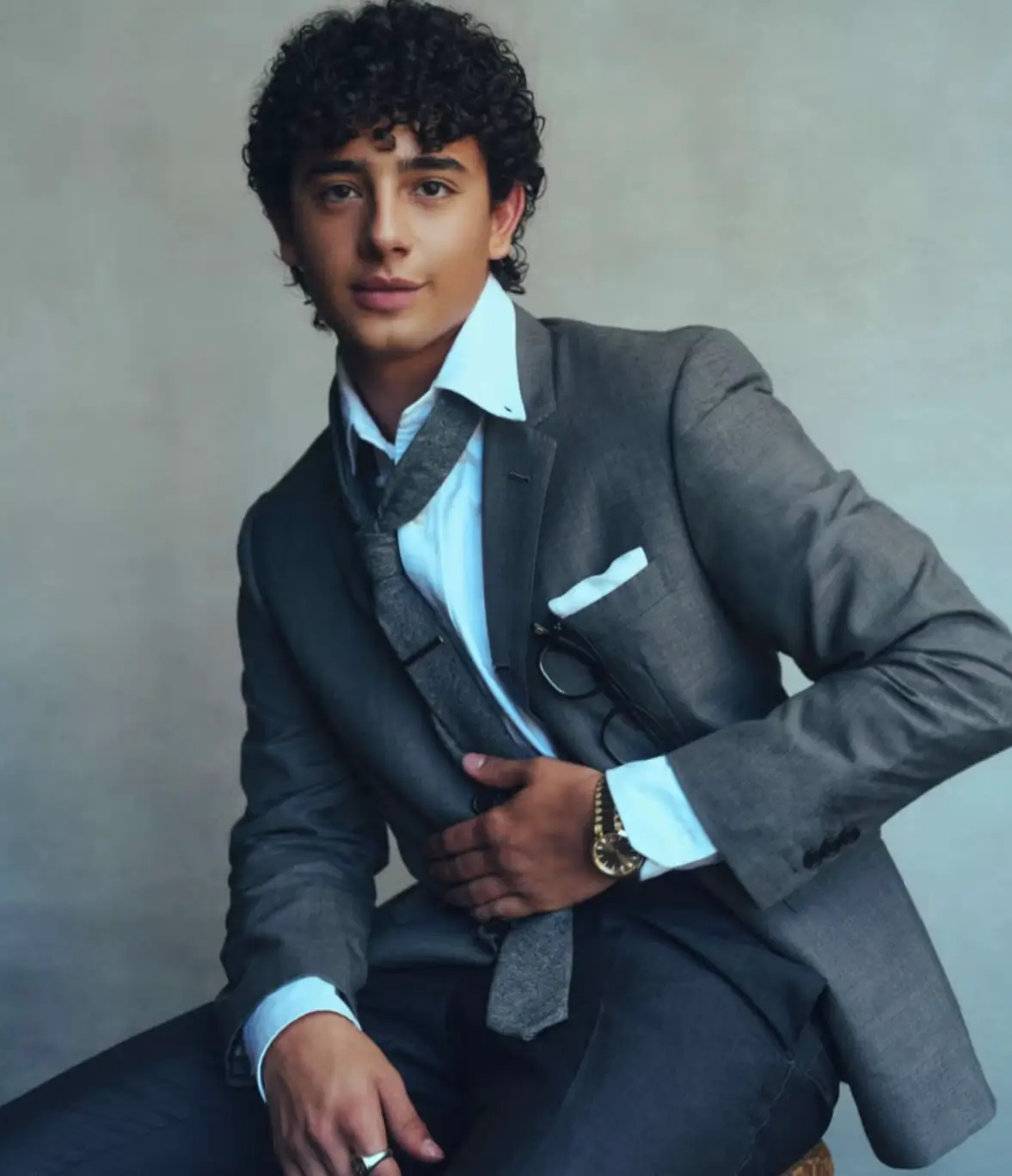 Studio portrait of a young person seated against a neutral backdrop, wearing a tailored gray suit with a white dress shirt and loosely knotted tie. Curly hair frames the face, while subtle accessories including a wristwatch, ring, and pocket square add polish to the relaxed yet sophisticated pose. Soft lighting and muted tones create a refined editorial fashion aesthetic