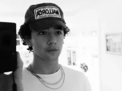 Black-and-white mirror selfie of a young person wearing a distressed baseball cap with a large front patch, a plain white T-shirt, and layered chain necklaces. Curly hair peeks out from beneath the cap, while the softly lit interior background remains out of focus, creating a casual, candid portrait with a minimalist aesthetic