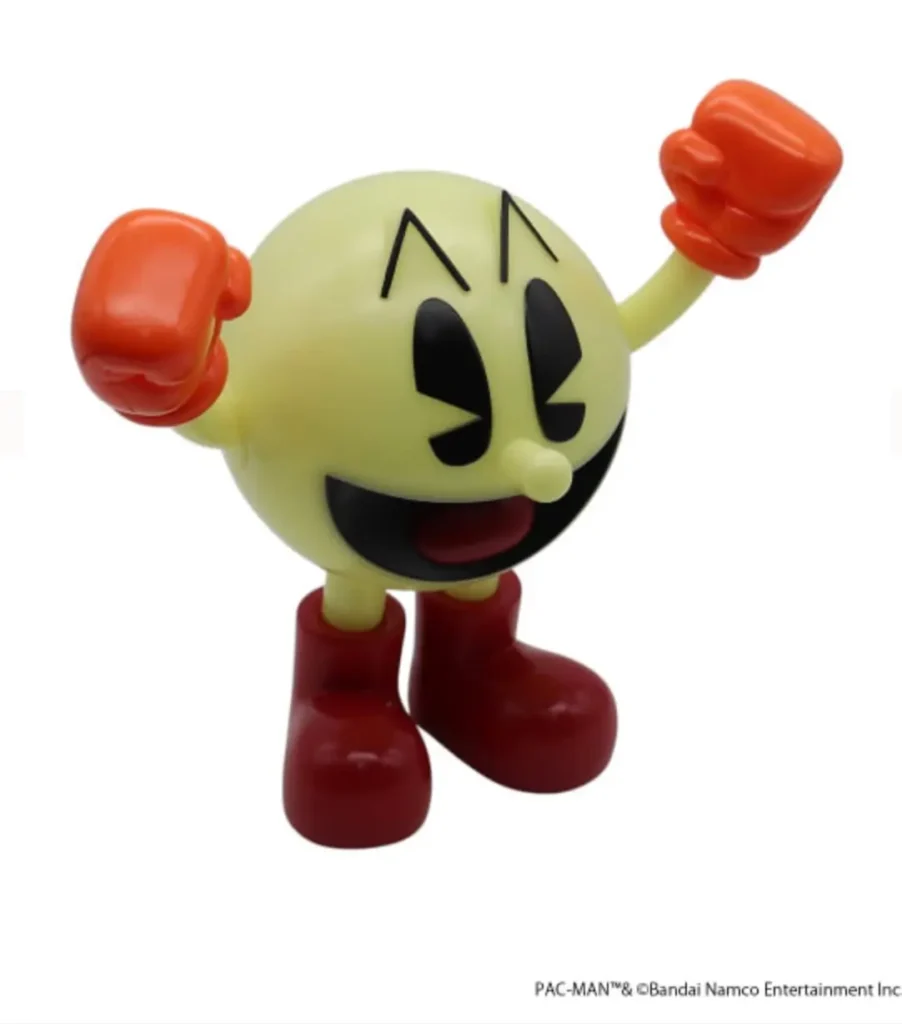 Three-quarter front view of the PAC-MAN figure posed with both gloved fists raised in celebration. The translucent yellow-green body, black facial features, and glossy red gloves and boots highlight the toy’s play arcade-inspired design