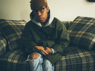Kenny Hoopla wearing a brown knit beanie, round black glasses, an olive-green jacket layered over a gray hoodie, and distressed light-wash jeans sits on a plaid couch in a softly lit room. The candid portrait features muted tones and a relaxed, introspective mood, emphasizing layered streetwear styling and a cozy indoor setting