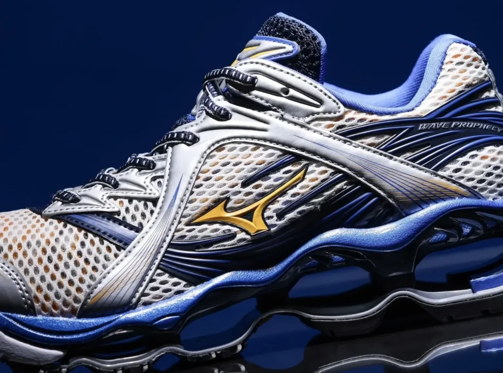 Detailed side-profile close-up of the Mizuno Wave Prophecy in a white, silver, blue, and gold colorway. The photograph highlights the metallic RunBird logo, open-mesh construction, layered overlays, and sculptural Wave sole against a rich blue background