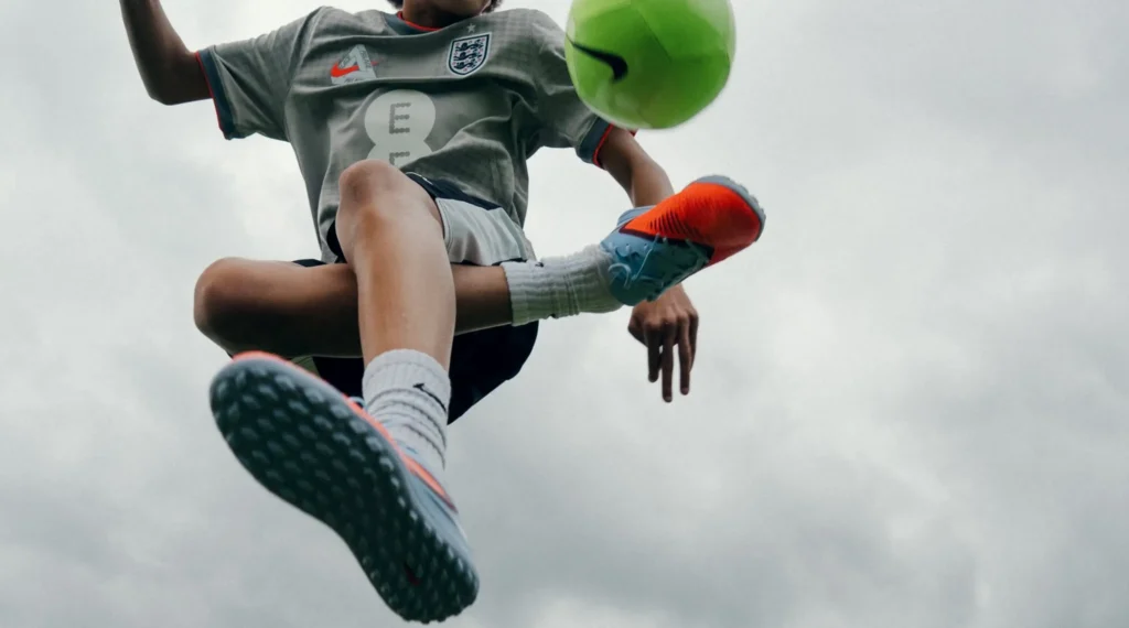 A young football player performs a skill move with a bright green Nike football suspended in midair. Captured from a dramatic low angle against a cloudy sky, the image highlights agility, control, and the excitement of youth football