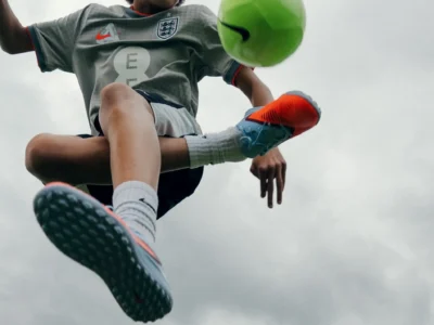 A young football player performs a skill move with a bright green Nike football suspended in midair. Captured from a dramatic low angle against a cloudy sky, the image highlights agility, control, and the excitement of youth football