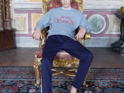 A fashion model relaxes in an ornate gilded chair inside a historic room with frescoed walls and a Persian-style rug. The outfit features a green baseball cap, gray graphic sweatshirt, navy trousers, and brown leather loafers, blending contemporary streetwear with a luxurious classical interior