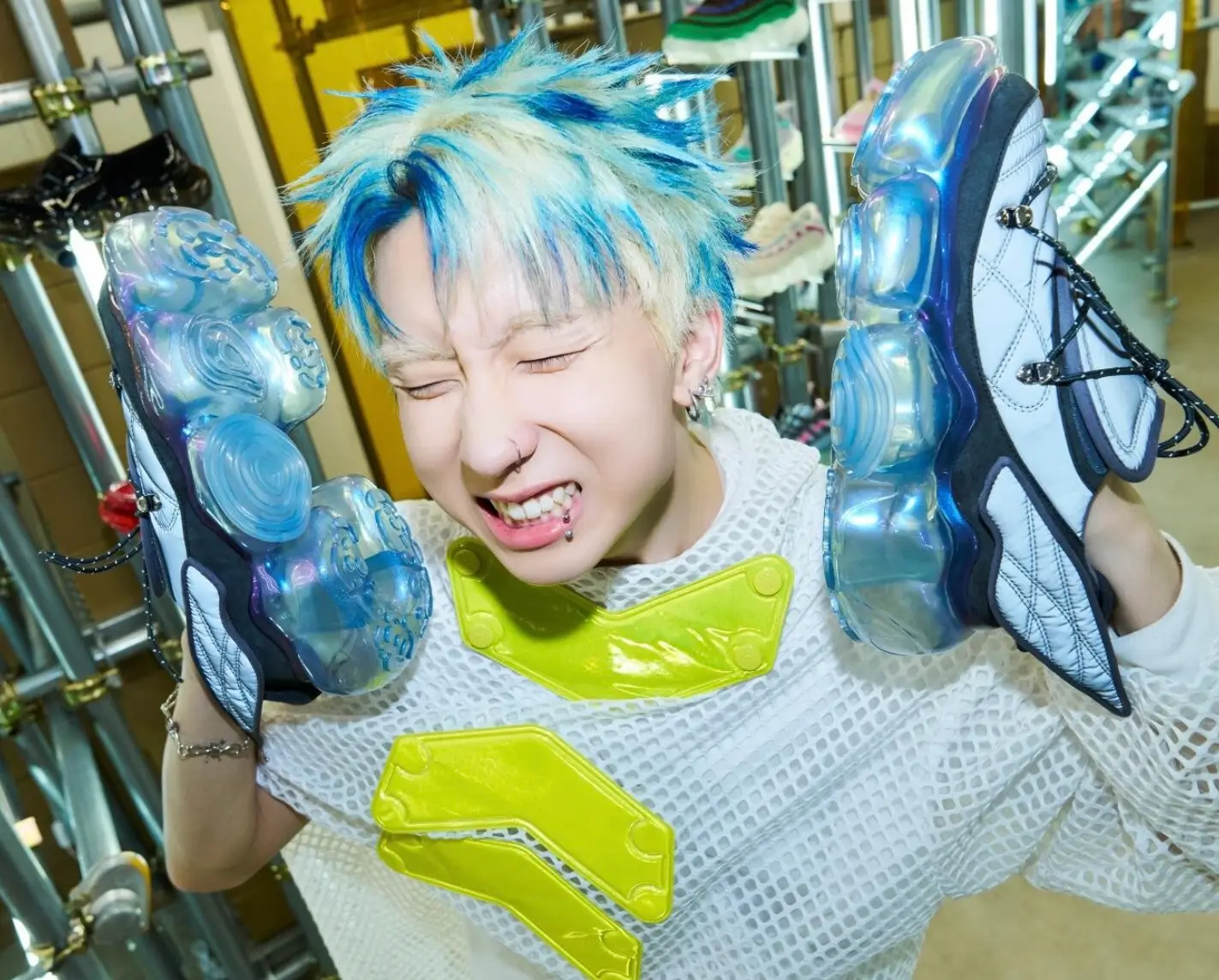 Person with blue-and-blonde spiky hair holding a pair of futuristic sneakers with translucent blue sculpted soles inside a sneaker boutique. Wearing a white mesh top with neon yellow accents, they smile with eyes closed while showcasing the bold footwear design.