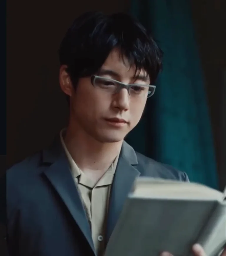 A young person with short black hair and translucent rectangular glasses reads an open hardcover book indoors. They wear a dark blue blazer over a light beige collared shirt with dark piping, creating a smart, understated look. Their expression is calm and focused as they look down at the pages. Soft natural light illuminates their face while the background remains dark and blurred, with a teal curtain adding subtle color contrast. The close-up portrait emphasizes concentration, quiet reflection, and a studious atmosphere