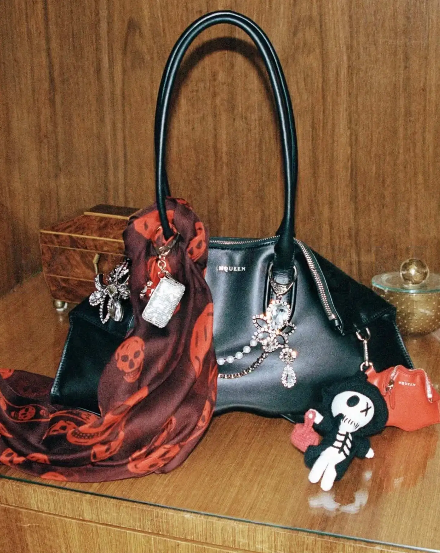 Alexander McQueen black leather handbag displayed on a wooden shelf, adorned with crystal charms, a red skull-print silk scarf, and playful character accessories that blend gothic luxury with contemporary personalization