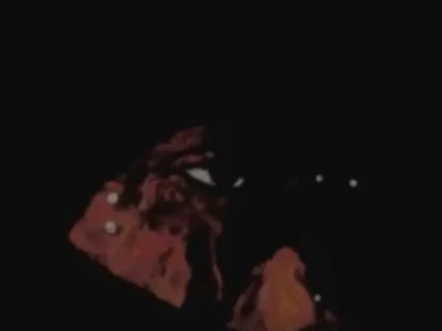 A dark, low-light image showing a sil figure with glowing white eyes emerging from near-total darkness, accented by deep red-orange highlights that create an eerie, atmospheric mood