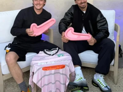 Two men sit on white lounge chairs holding oversized pink Nike Swoosh props, with a pink football jersey displayed between them. A soccer ball rests on the floor in front, while one man wears colorful football boots and the other sports Nike Air Max 95 sneakers, creating a casual sportswear and football culture scene