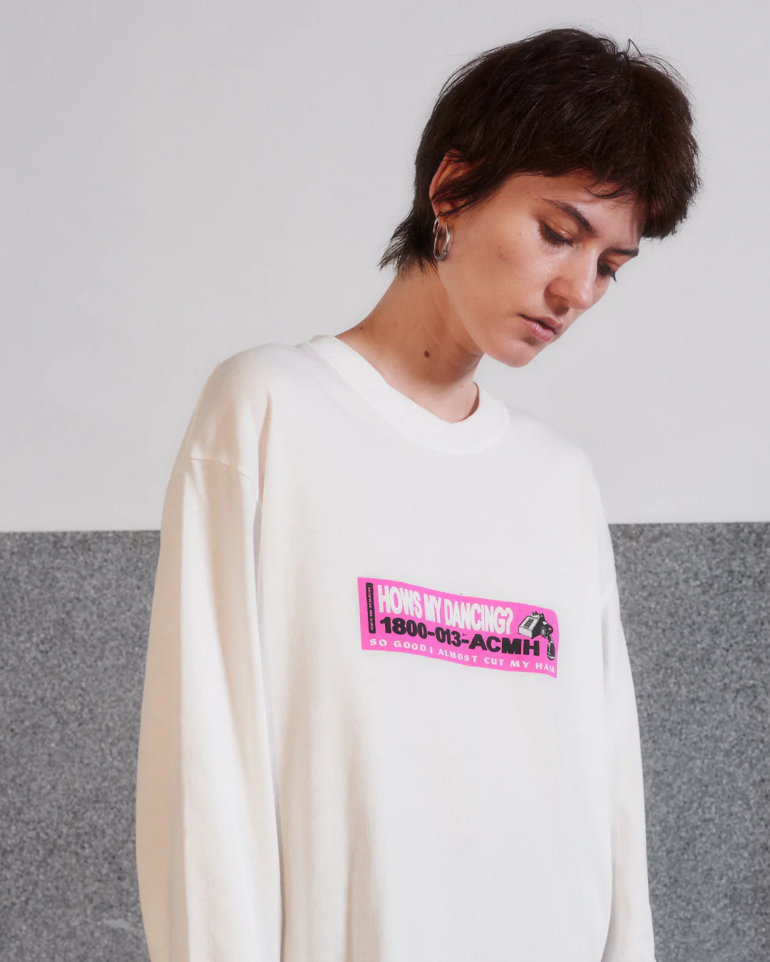 Model wearing an oversized white sweatshirt with a bright pink graphic logo on the chest, photographed against a minimalist gray backdrop