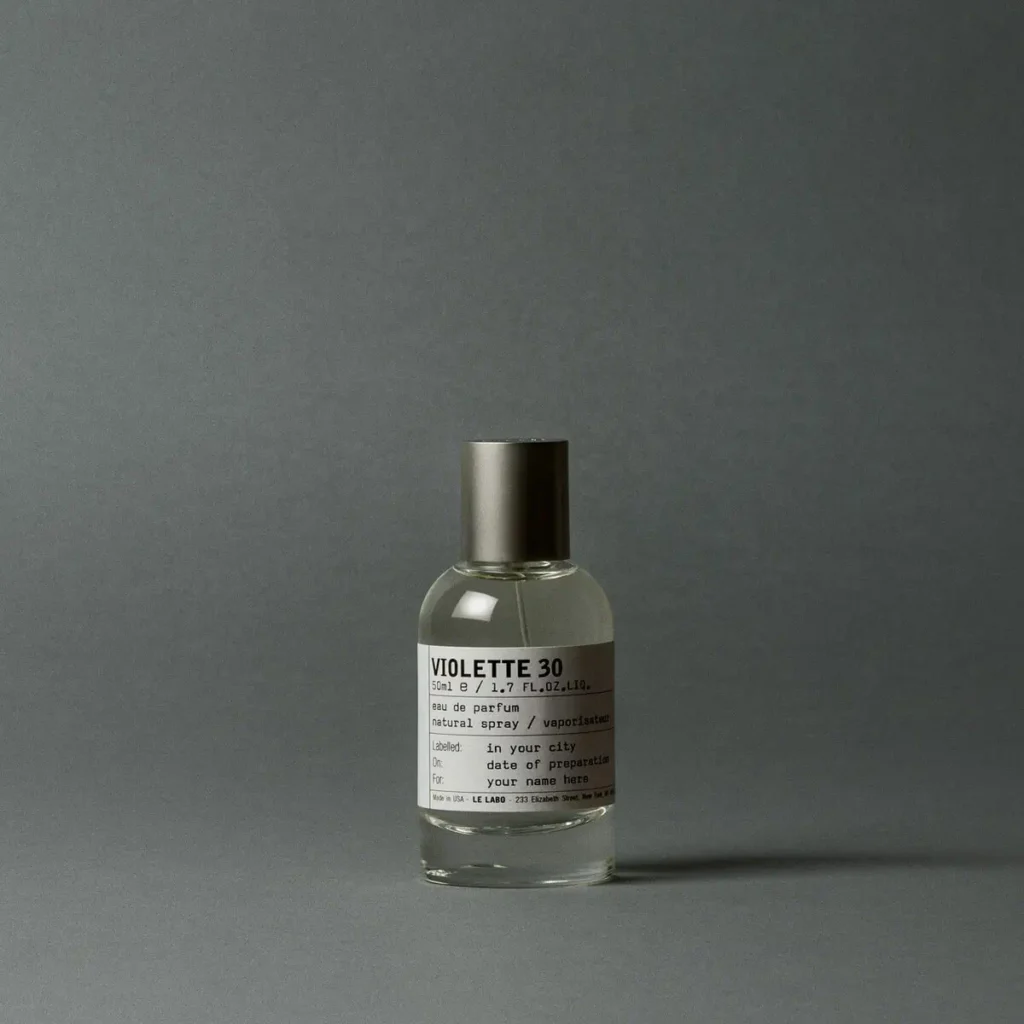 Le Labo Violette 30 Eau de Parfum bottle photographed against a minimalist charcoal-gray backdrop, showcasing the fragrance’s clear glass flacon, dark metallic cap, and signature laboratory-inspired label