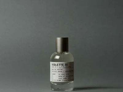 Le Labo Violette 30 Eau de Parfum bottle photographed against a minimalist charcoal-gray backdrop, showcasing the fragrance’s clear glass flacon, dark metallic cap, and signature laboratory-inspired label