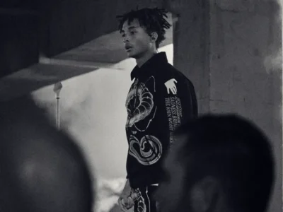 Black-and-white editorial photo of a Jaden Smith wearing a dark graphic jacket under a concrete overpass, with blurred figures in the foreground creating a moody, urban atmosphere