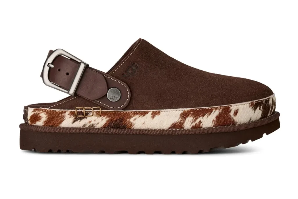 Side profile of a UGG suede clog in dark brown with an adjustable leather heel strap and silver-tone buckle. A cow-print calf hair wrap surrounds the midsole, contrasting with the soft suede upper and chunky rubber platform