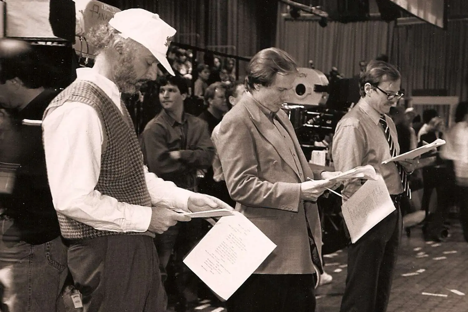 Black-and-white behind-the-scenes photograph of a television production set, showing several crew members and creatives standing side by side while reviewing script pages. Large studio cameras, lighting equipment, and production staff fill the background, capturing the atmosphere of a busy rehearsal or film session